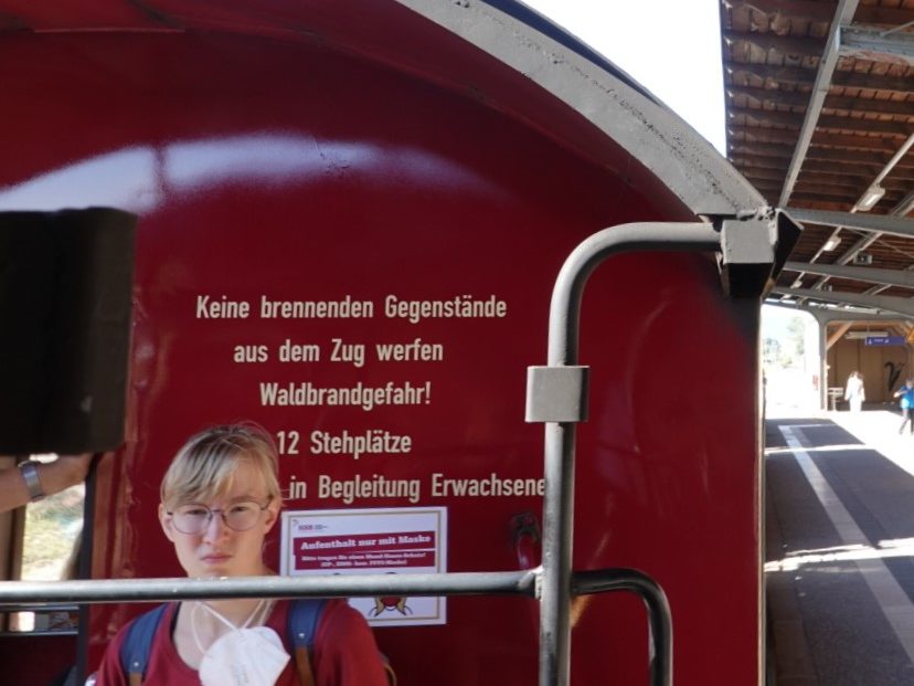 A narrow gauge train car with a red exterior displaying safety instructions in German, and a young person standing in front of it at a train station.