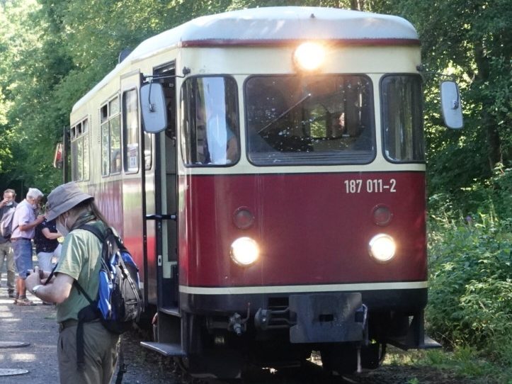 A Harz narrow gauge railway diesel car train unit No. 187-011-2 with a red and cream exterior is parked along a tree-lined track, with passengers gathered nearby.
