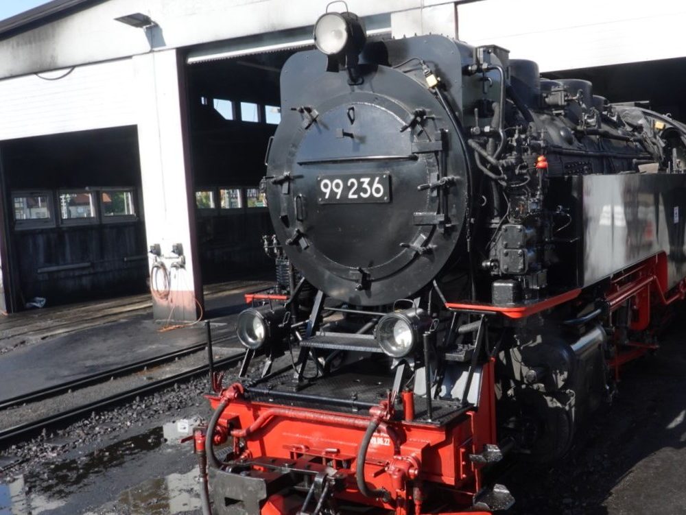 A close-up of a Harz narrow gauge railway steam locomotive No. 99-236 standing at Wernigerode, showcasing its black and red exterior. The train is positioned inside a train shed, surrounded by industrial elements.