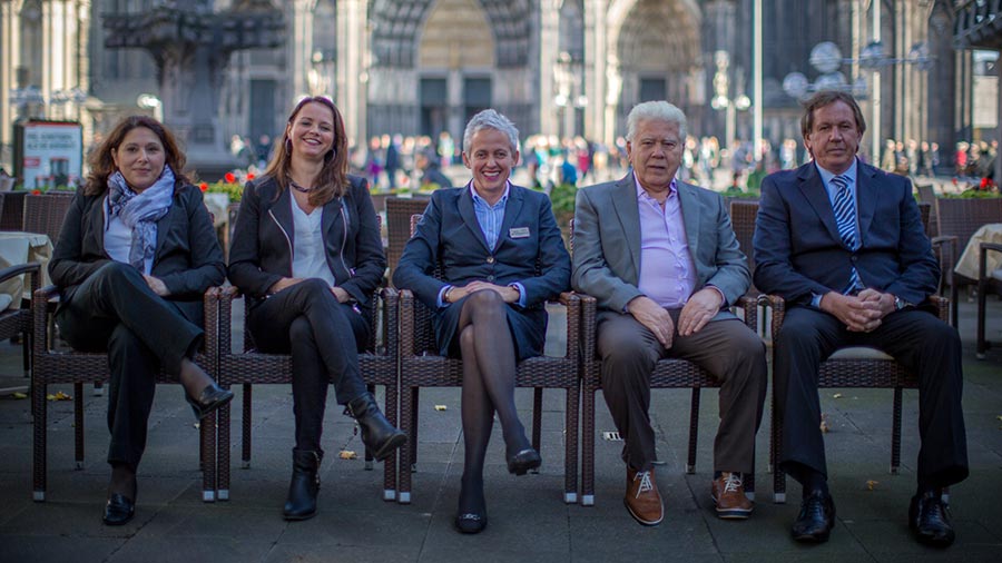 Five staff from the Cafe Reichard seated on chairs with Cologne Cathedral in the background.