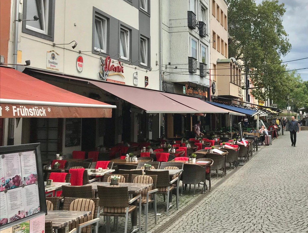 Outdoor dining area of a restaurant in Unter Kaster, Old Town, Cologne with tables and red seat cushions along a cobblestone street.