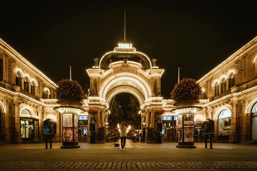 Night view of the illuminated main entrance to Tivoli Gardens in Copenhagen, with decorative lights and flowers. Tivoli Gardens forms the starting point for this self guided walk through central Copenhagen 