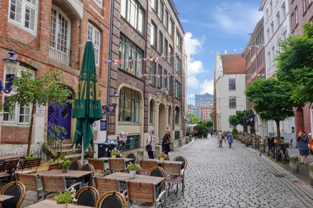 A view of Deichstrabe in Hamburg featuring cobblestone pavement, outdoor dining areas with tables and chairs, and colorful strings of flags decorating the space. Historic buildings line one side of the street while red brick and modern architecture makes up the other side of the street. In the distance there are a few pedestrians strolling along and a few bicycles are parked nearby.