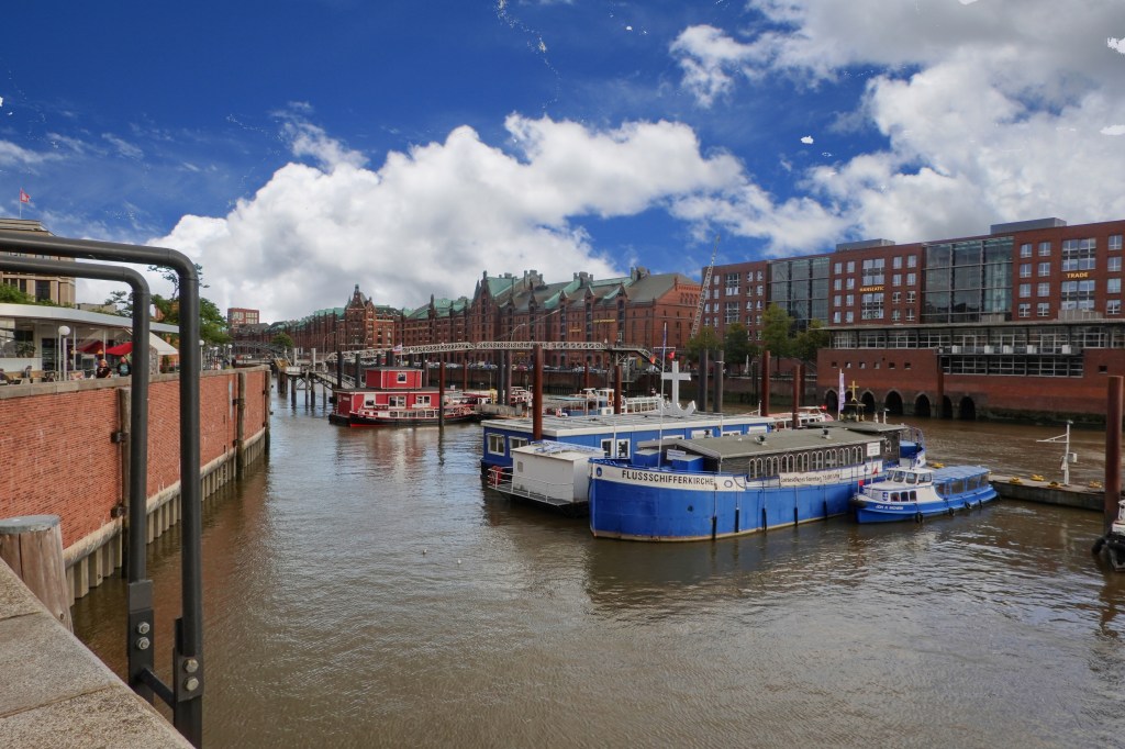 A scenic view of the Norderelbe in Hamburg, featuring various boats docked along the water, surrounded by the brick buildings of the Speicherstadt and a bright blue sky with clouds.