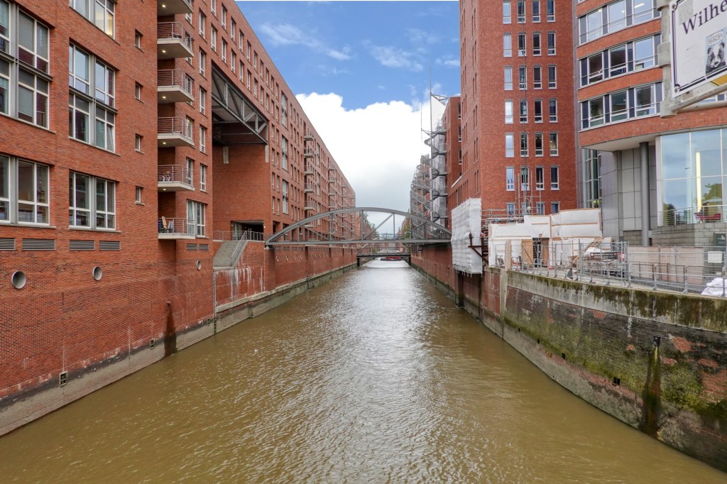 View of the Sankt Annenfleet waterway in Hamburg surrounded by the red brick buildings of the Speicherstadt. In the distance there is a small bridge spanning the waters of the Sankt Annenfleet