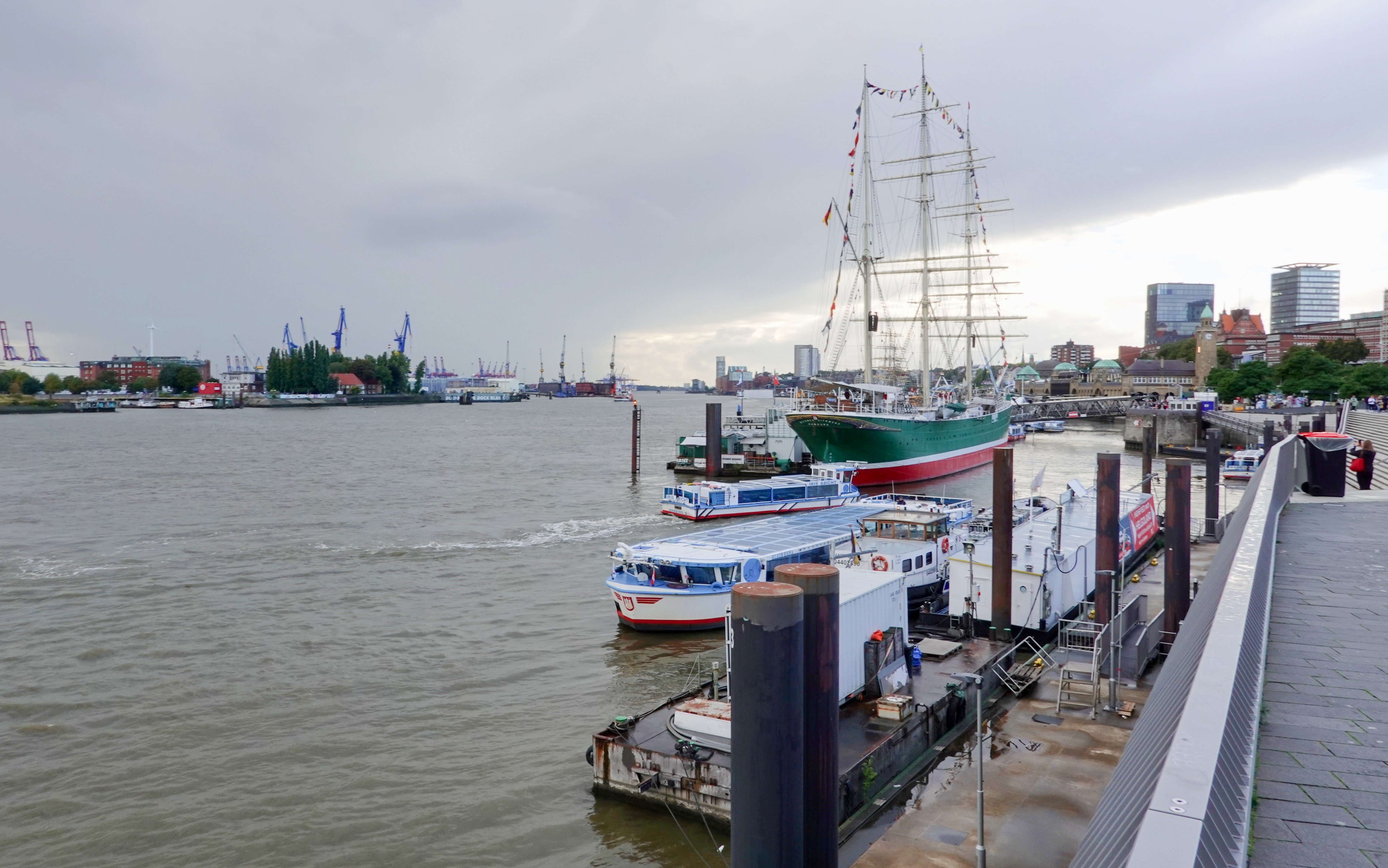 A view of the River Elbe in Hamburg featuring various boats docked at the quayside with the tall sailing ship Rickmer Rickmers in the foreground and cranes of Hamburg port and modern buildings in the background all under a cloudy sky.