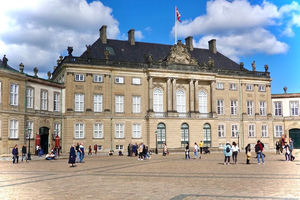 Exterior view of Amalienborg Palace in Copenhagen with visitors in the courtyard and blue sky with clouds. The Amalienborg Palace forms another key attraction on this self guided walk route through central Copenhagen 

