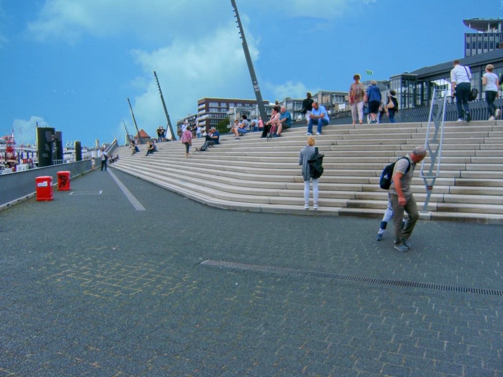 View of the bustling Jan Fedder Promenade in Hamburg featuring people sitting on steps and enjoying the view of the waterfront.
