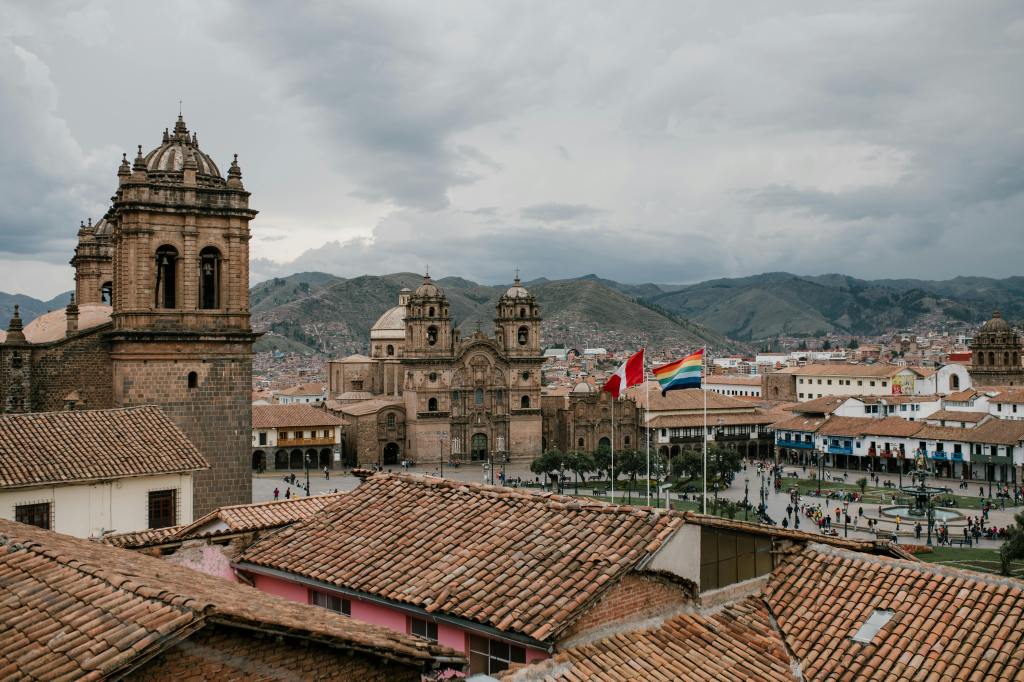 A panoramic view of Cusco, Peru, showcasing the historic architecture including colonial churches and tiled roofs, with the Andes mountains in the background under a cloudy sky. Cusco is an ideal centre for budget travellers, with cheap hostel and hotels and affordable eateries