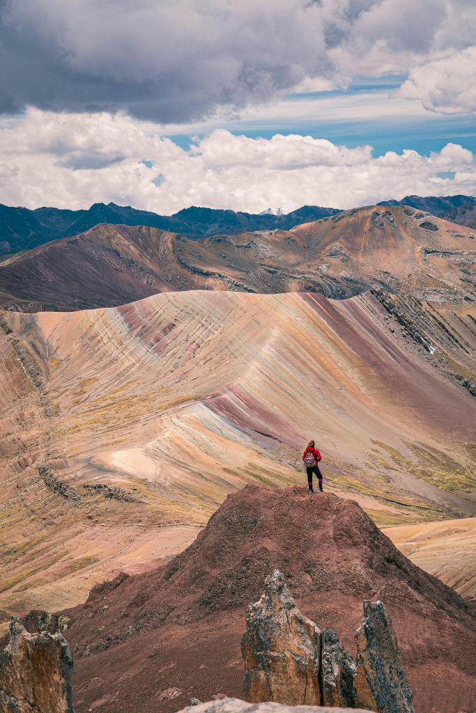 A hiker standing on a rocky peak overlooking the colorful hills of the Andes mountains in Peru under a cloudy sky. This terrain is ideal for backpacking, hiking and travelling on a budget