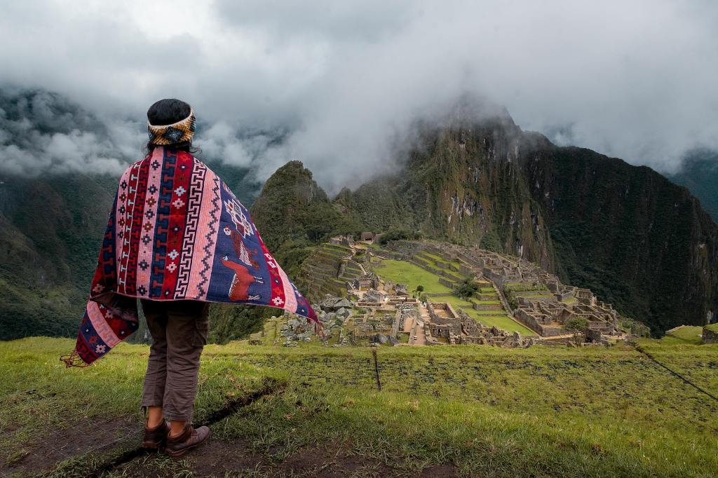 A person wearing a colorful traditional poncho stands overlooking the ancient Inca citadel of Machu Picchu, surrounded by lush green mountains and a cloudy sky.
