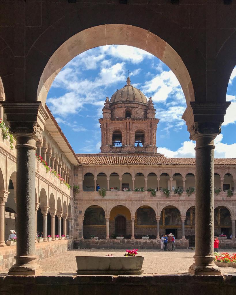 View of a historic courtyard of the Coricancha in Cusco, Peru. The image is framed by arches, showcasing colonial architecture and a bell tower against a blue sky.