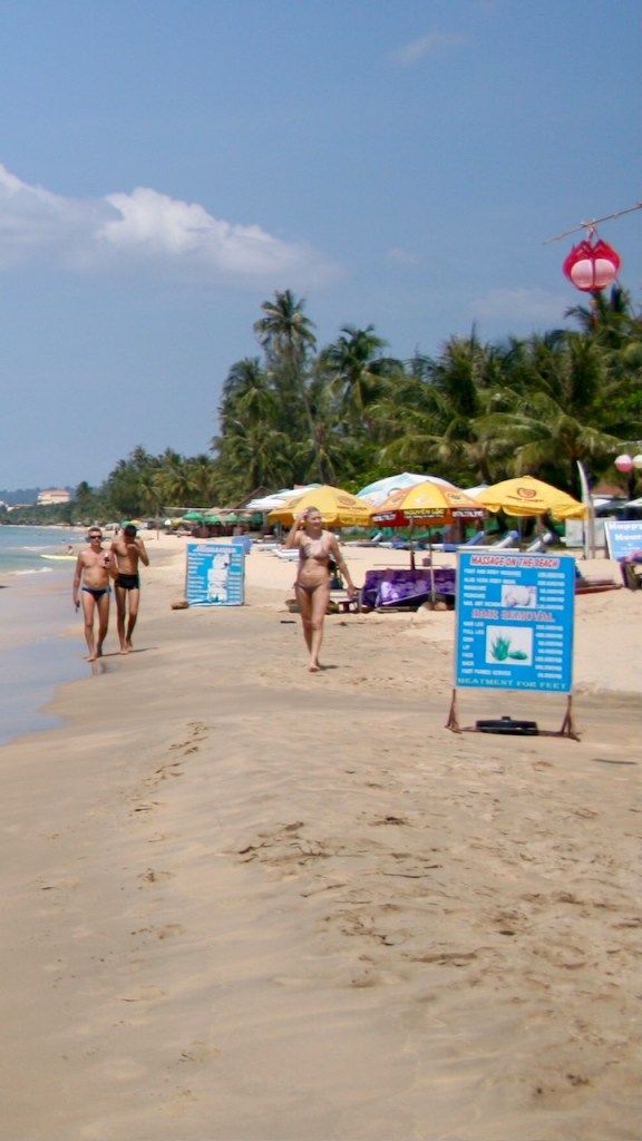 The photo shows people walking on the beach at Bai Truang Beach. Phu Quoc, Vietnam. The photo includes a sandy beach, sun umbrellas and beach merchant sign boards