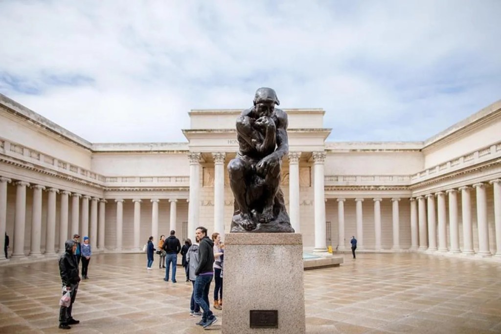 This image shows the Court of Honor within the Legion of Honor in San Francisco. In the forefront of the picture, set on a concrete plinth, is an original bronze cast of Auguste Rodin's 'The Thinker'. This is one of 29 original casts located around the world. Behind this original cast is the columnated courtyard area of the Legion of Honor. Some people are shown looking around the courtyard area. Above this scene is a cloudy blue sky.