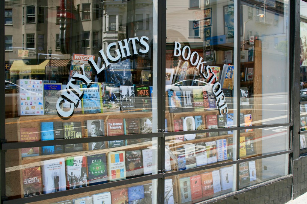 This image shows the window of the City Lights Bookstore. The words 'City Lights Bookstore' are stencilled onto the window in curved semi circle and the shop behind contains a display of books. Reflections of the street can be seen in the window. These reflections show images of the surrounding buildings and the cars using the street.