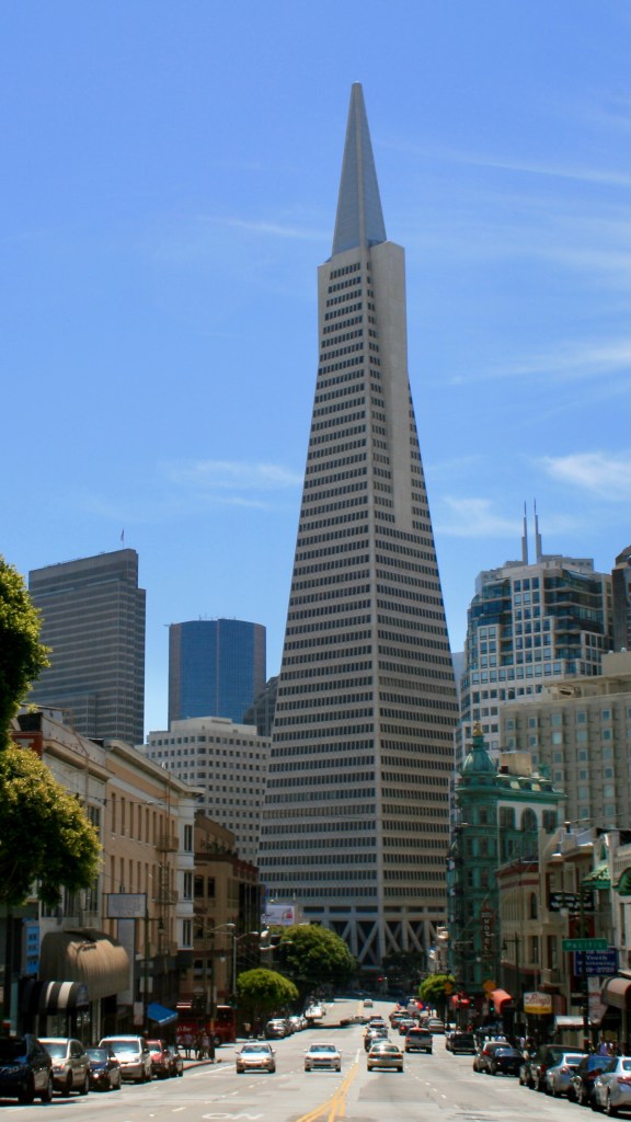 This image shows the TransAmerica Pyramid Building in North Beach, San Francisco as viewed from the intersection of Columbus and Kearney. The Pyramid Building is shown as a monolith, blocking off the southeastern end of Columbus and towering above its surroundings. In the foreground the traffic on Columbus is dwarfed by the scale of the building, as are the other tall buildings shown in the background of the picture.  The top half of the picture shows solely the tapering pyramid and a pale blue sky, The overall impression of the scene is that of a powerful space age rocket sitting on its launchpad in the middle of a city.