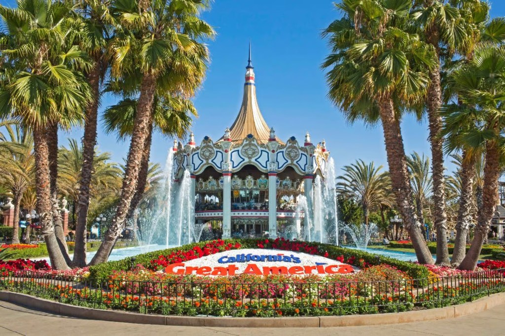 This image shows. in the foreground, a flower bed, fringed with palm trees. At the centre of the flower bed is a display with the wording 'California's Great America'. Behind the flower bed there is a water feature with water fountains and a fairground attraction at the far end. This scene is set against the background  of a blue sky