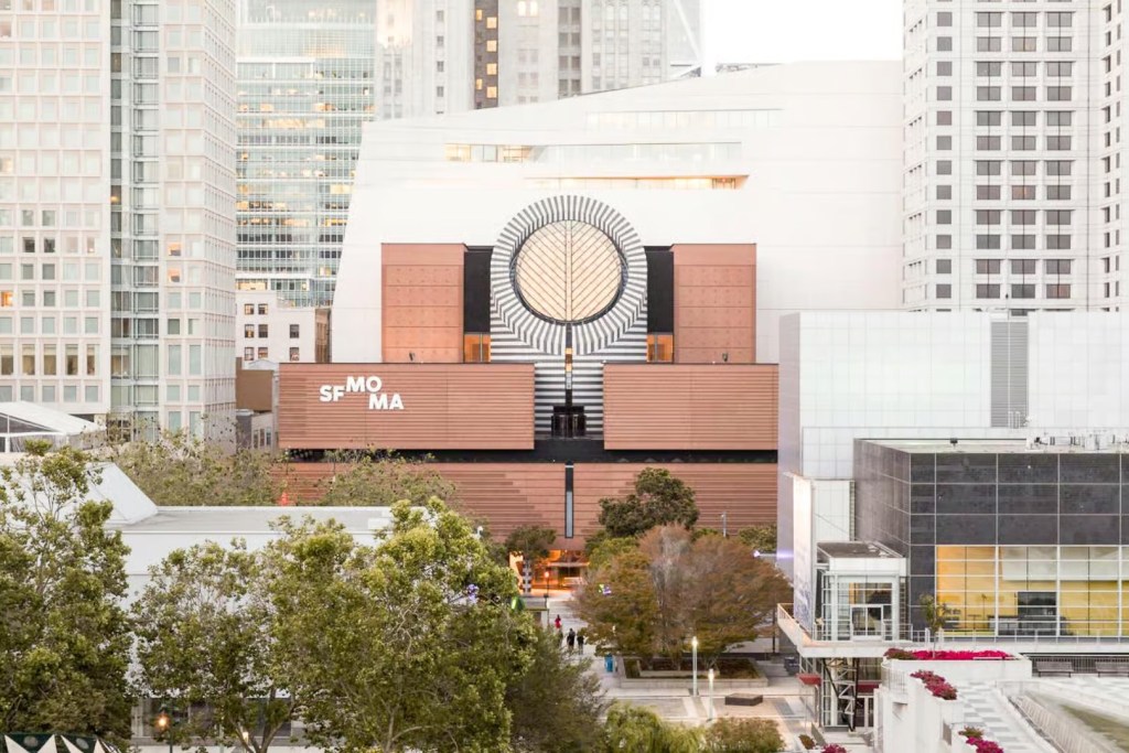 This photo shows the exterior of the San Francisco Museum of Modern Art, San Francisco, California. The building is of a modern design, being generally white in colour with a brickwork entrance area. The word 'SF MO MA' is spelt out in an irregular pattern on the brickwork above the entrance area. Also atop this brickwork entrance structure there is additionally a large circular gold and white motif, set within a black and white ring and set upon a black and white horizontally striped podium.   