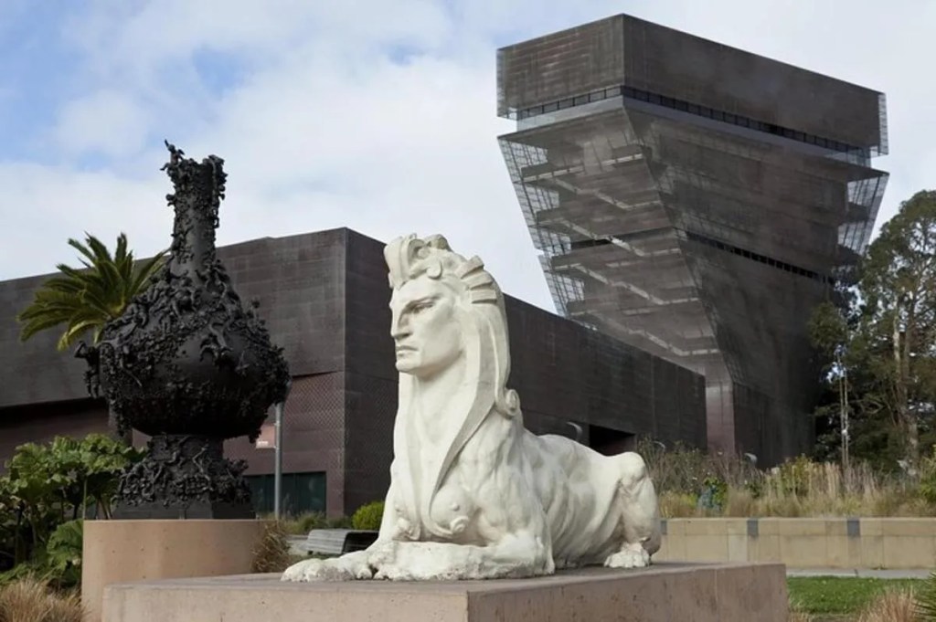 This image shows the outside of the de Young Museum in San Francisco. In the foreground there is an Egyptian Sphinx on a plinth and it the background there is the futuristic structure of the museum itself. Above it all there is a light blue cloudy sky