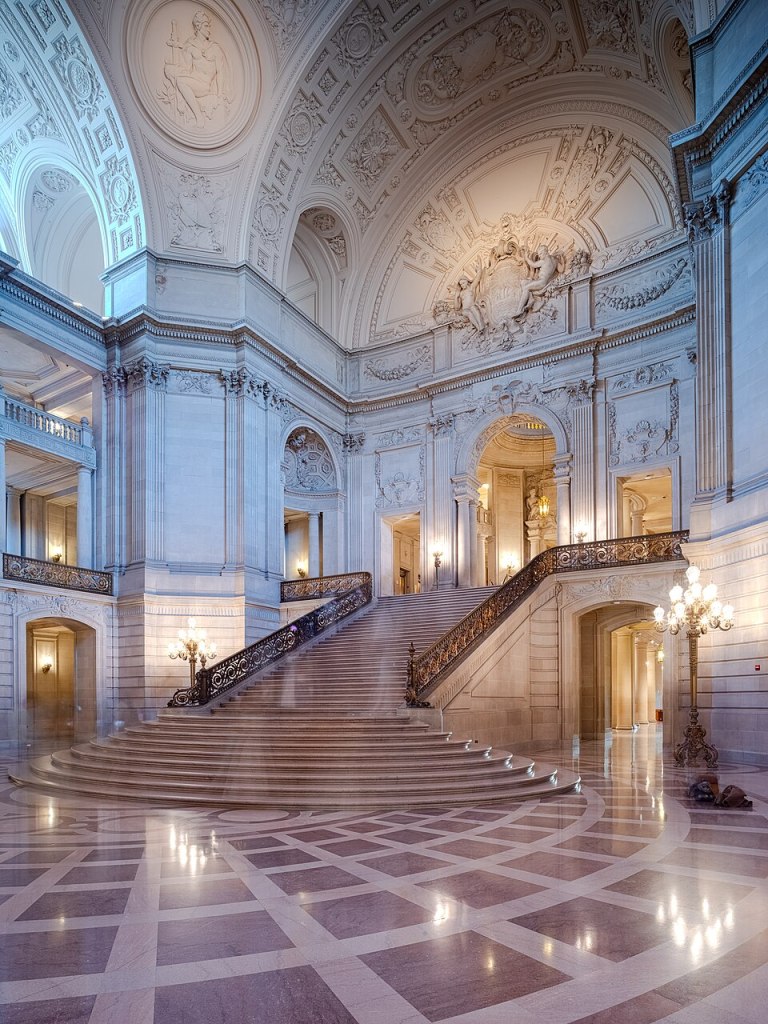This image shows the interior of  San Francisco's City Hall. In the image the marble flooring and sweeping staircase from the entrance area up to the first floor area can be seen. Also the detailing  of the walls and the ceiling is also visible.