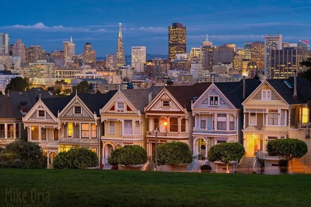 This image shows the Painted Ladies. These are a row of seven Queen Anne Victorian houses located on the north side of Alamo Square, San Francisco. The photo has been taken in the early evening from within Alamo Square and shows the houses against a backdrop of the modern City's skyscrapers and a darkening evening sky. Among the skyline, the shapes of the Pyramid Building and Coit Tower can be seen Because of the time of day, the windows of the Painted Ladies houses are illuminated by internal house lighting. 