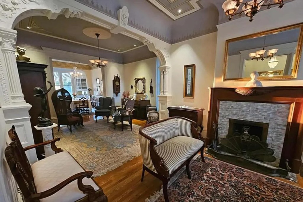 This image shows the interior of one of the Painted Ladies houses. The room and its furnishings are set out with period decor that match the age of the building itself. In the foreground are two lounge chairs and a fireplace and in the background there is more period furniture and a full length window opening onto what looks like a garden.