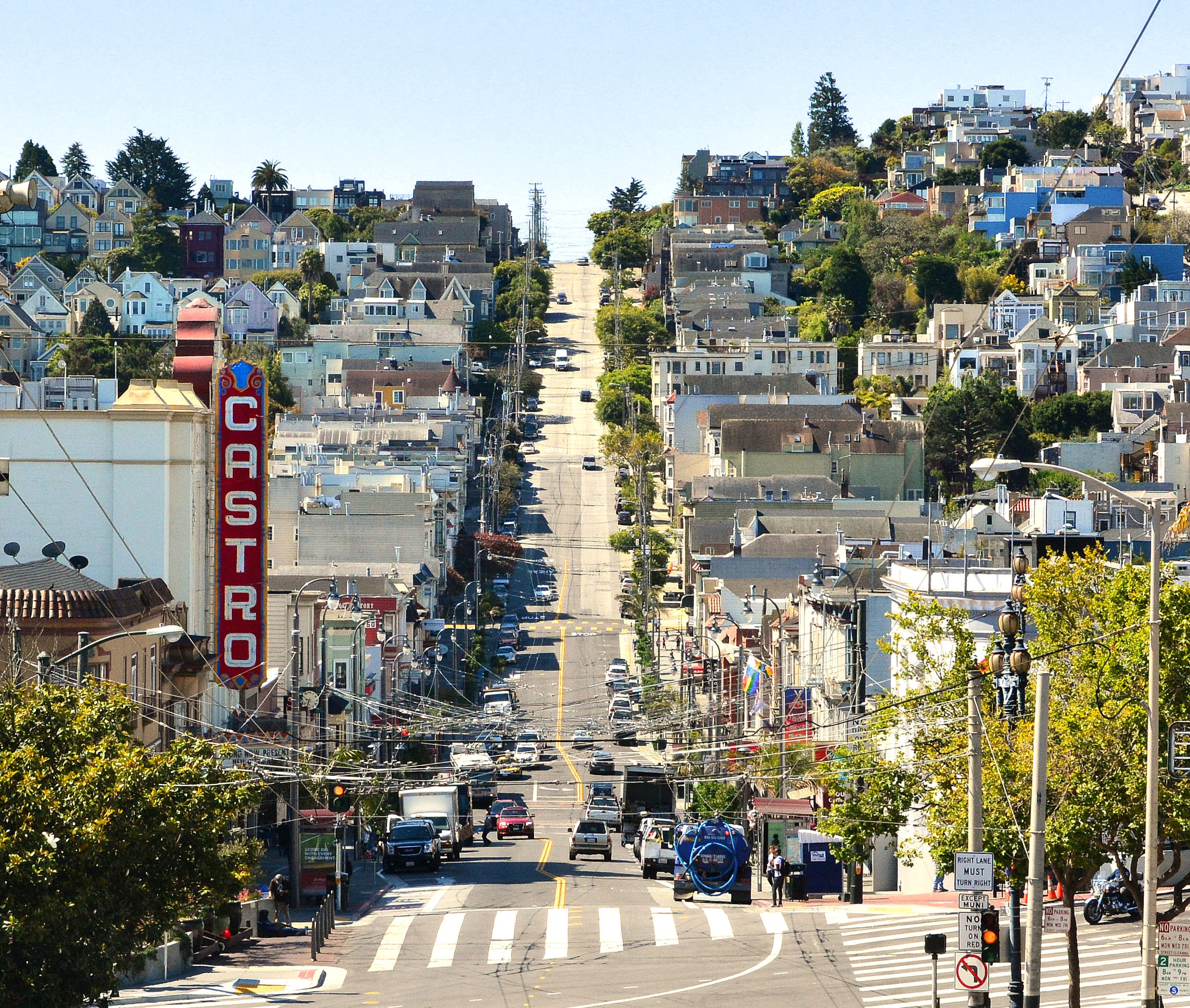 This image shows a front-on view of Castro Street, San Francisco, looking southwards from its intersection with Market Street. On the left hand side of the street, in the foreground, can be seen the distinctive white lettering on red background 'Castro' sign, advertising the Castro Theatre. From the intersection Castro Street is seen initially descends towards 18th and 19th Street before climbing up to Billy Goat Hill in the far distance.  In the image the roadway of Castro Street is lightly trafficked. On either side of the road the footways are lined with greenery from shrubs / trees and the surrounding buildings are generally flat roofed two or three storey apartments. The top quarter of the picture, above the buildings, is occupied by a watery light blue sky.