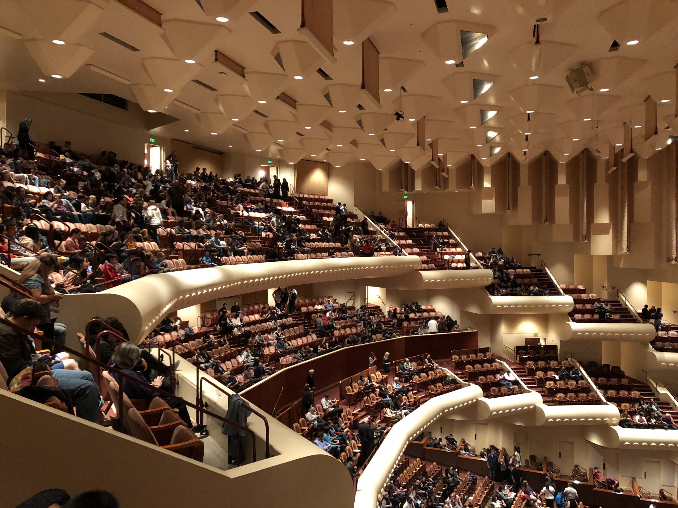 This image shows the interior of the Davies Symphony Hall in San Francisco. The image has been taken from high up in the building, to one side of the stage. It shows roofing area together with the arrangement of circle area seats, tiered steeply downwards towards the stage below. The stall area seating arrangements can also be seen in the bottom right of the photo.