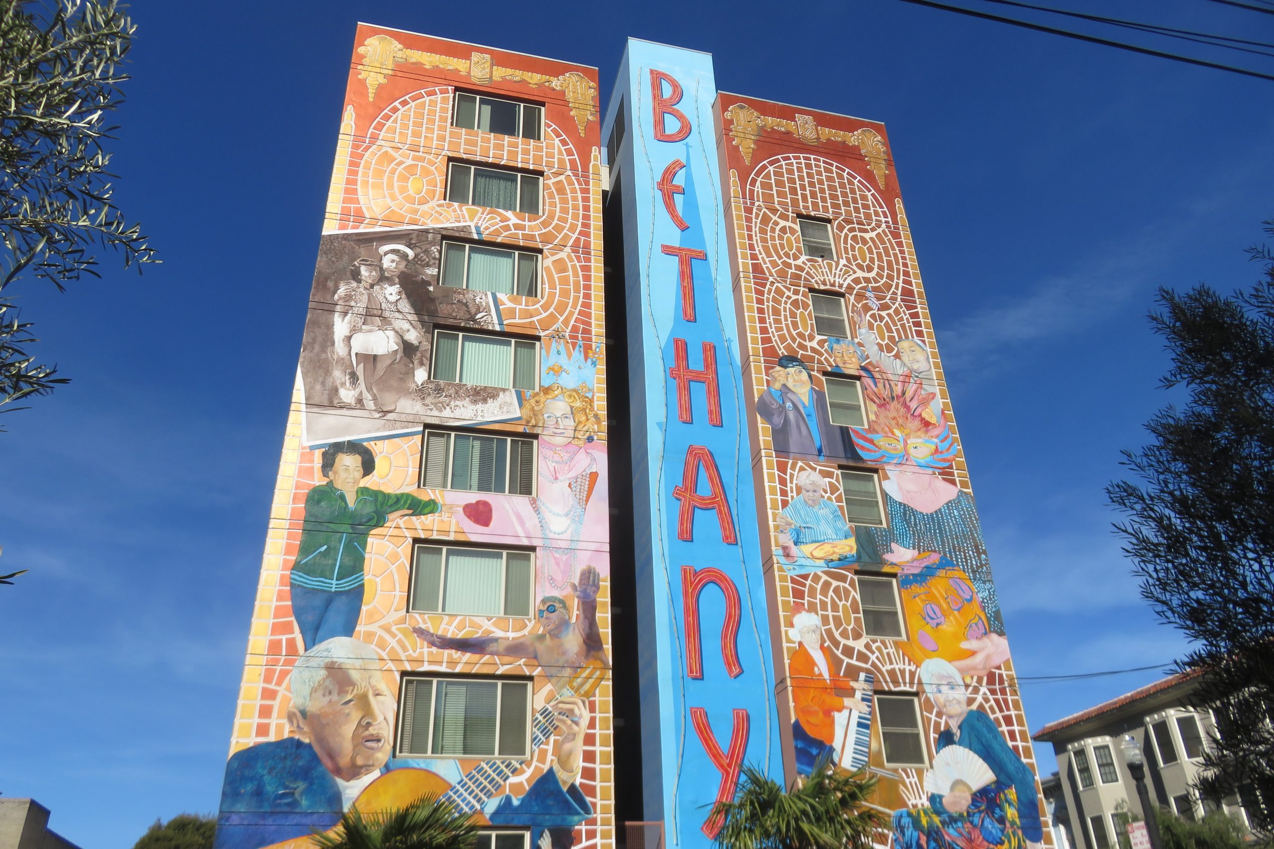 This image shows the full height 'Salud!' mural at the Bethany Centre in Mission, San Francisco.  The mural is eight storeys high and 75ft wide, covering the south wall of the building. On either side of what appears to be a central lift shaft or stairwell, the mural depicts scenes based around the lives of the senior residents that live within the Bethany Center. The base colour for this part of the mural is mixture of yellow and orange, with the residents being depicted in contrasting colours. In the central part of the mural the stairwell / lift shaft has been painted a sky blue colour, with a 'shadow' of the nearby church being imposed on top with a darker shade of blue colour. This base colouration has then been overlaid in red with the word ~Bethany', This word has been painted in 10ft high letters painted vertically from top to bottom. This image is then set off in the photograph against a dark blue sky and bordered by trees. Also, due to the choice of photographic lens or the chosen viewpoint, the photo, gives the mural an appearance of tapering towards the sky.