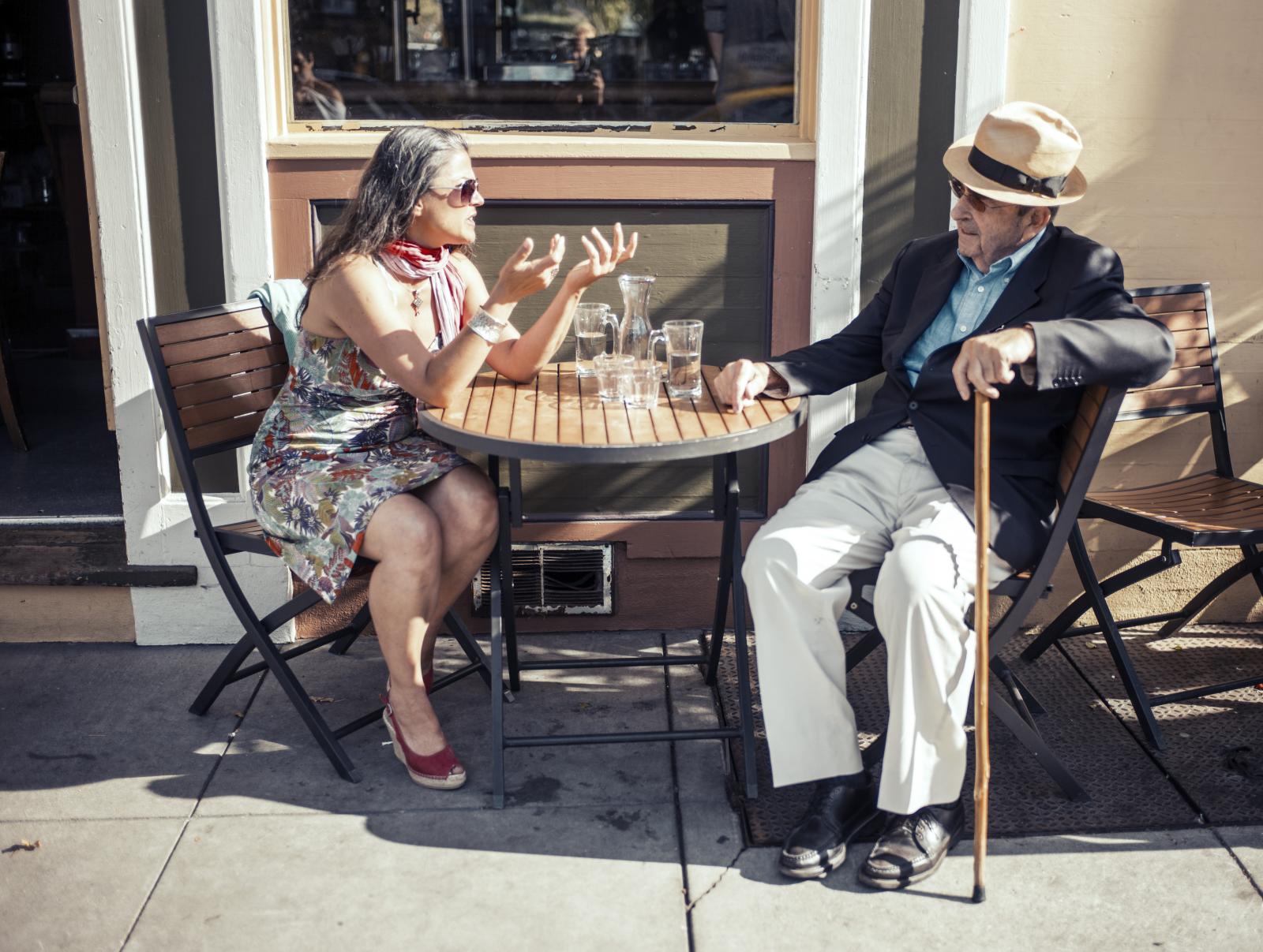 This image shows a well dressed older Italian looking gentleman and an older well dressed lady sitting at a round slatted wooden table at a street cafe / bar in North Beach. The man is wearing white trousers, a black blazer, blue shirt and black shoes and has a walking cane in his hand a straw hat on his head. The lady is wearing a printed pink and grey coloured summer dress and  has a matching silk looking scarf tied round her neck.  There are two glasses and an empty carafe on the table.