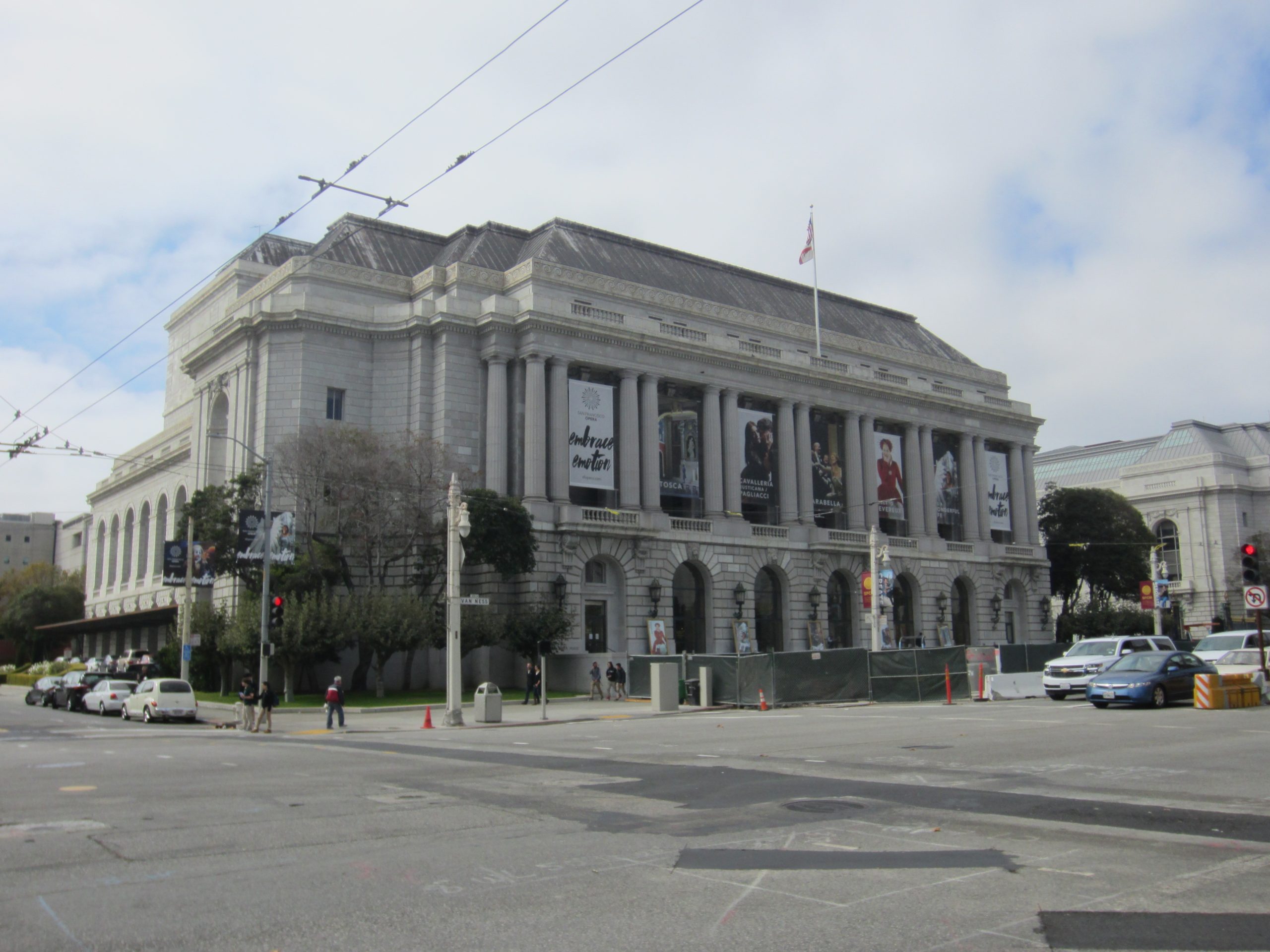 This image shows the exterior of the War Memorial Opera House in San Francisco.. The front of the building is viewed from the street. There are trolley bus wires shown in the foreground, a flag is flying above the building and the sky is primarily cloudy.