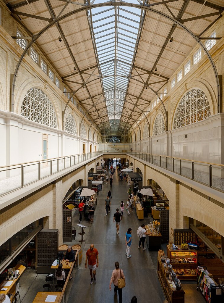 This image shows the interior south wing of the San Francisco Ferry Building at Fisherman's Wharf. The picture has been taken from the upper gallery level of the building. At ground level, below the gallery,  the picture shows people moving between the various retail stalls that occupy the ground floor.  At upper level the building has a utilitarian feel to it, with galleried walkways running down either side of the wing and a glass roof being present above, supported on a system of steel trusses. The scene is reminiscent of a Victorian market hall or even a Victorian prison building.