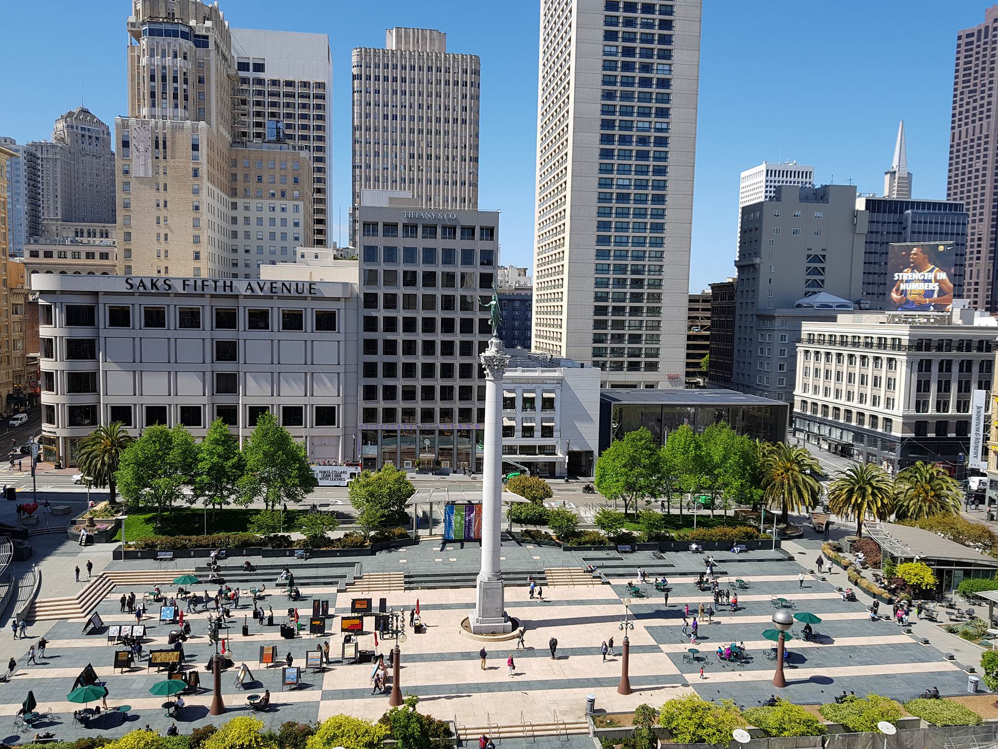 This image shows a view of Union Square, taken from an elevated position. The photo shows the main paved area of the Square with people going about their daily activities. In the background are the high rise buildings that surround the Square and all of this sits under a blue sky. In the centre of photo, in Union Square, sat on a 85 ft column, is the statue of Nike, the Greek goddess of victory. The column and the statue are dedicated to Admiral George Dewey. in celebration of his victory in 1898 over the Spanish at the Battle of Manila Bay during the Spanish - American War.. The statue and column, combined, are known as the Dewey Monument.